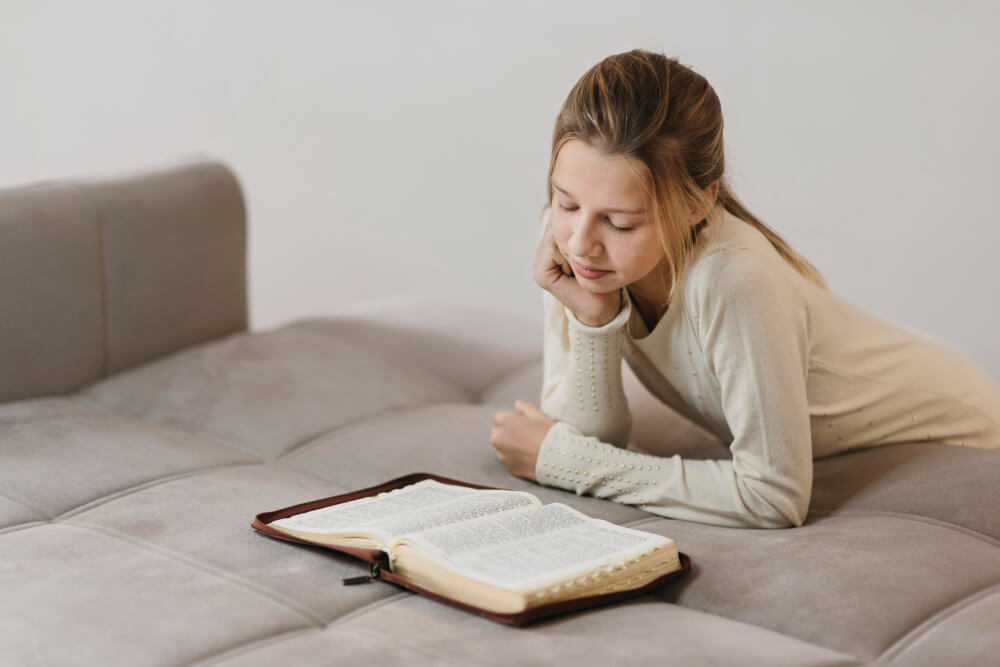 Menina deitada em sofá lendo Bíblia aberta, demonstrando momentos pessoais de leitura da Bíblia para crianças.