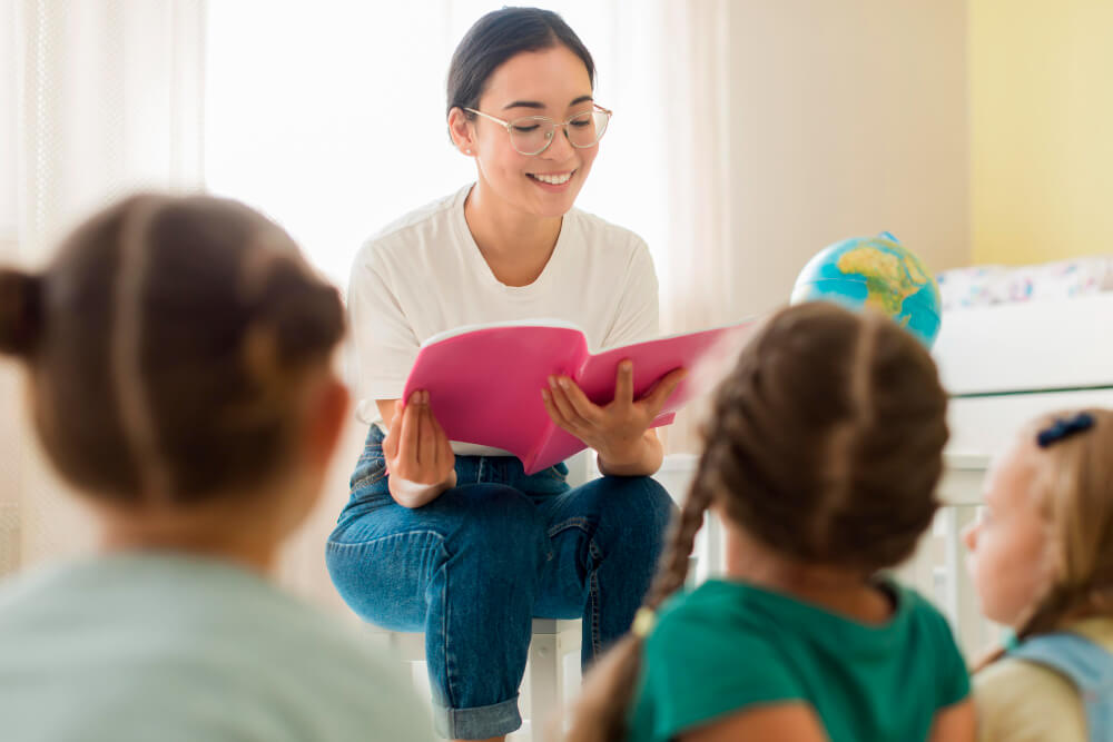 Mulher jovem lendo um livro rosa para um grupo de crianças pequenas sentadas à sua frente em uma sala iluminada, representando uma líder do ministério infantil conduzindo uma atividade educativa.