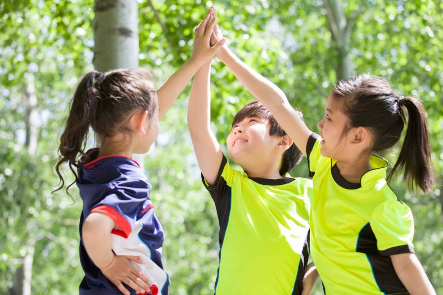Três crianças com uniformes esportivos fazendo high-five ao ar livre, celebrando como se estivessem vivendo a copa do mundo para crianças