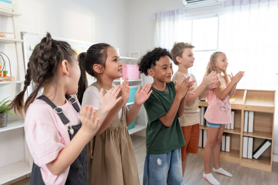 Grupo de cinco crianças de pé em uma sala, batendo palmas juntas durante uma atividade do dia do pastor no ministério infantil